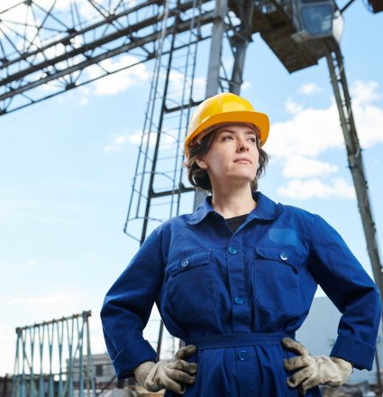 Waist up portrait of empowered woman working at construction site posing against sky with tower crane in background, copy space