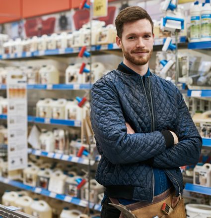 Confident handyman in a hardware store standing amongst the racks of products with folded arms smiling at the camera