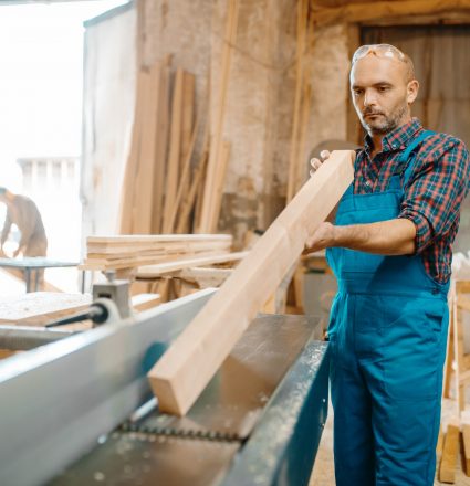 Carpenter with board near plane machine, woodworking, lumber industry, carpentry. Wood processing on furniture factory, production of products of natural materials