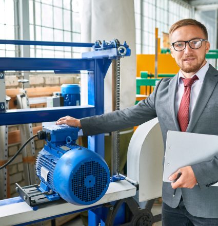 Portrait of successful salesman wearing suit posing looking at camera, standing by machine tools in industrial showroom