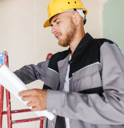 Engineer in work clothes and yellow hardhat dreamily looking on Engineer in work clothes and yellow hardhat dreamily looking on plan of new apartments leaning on red ladder at work